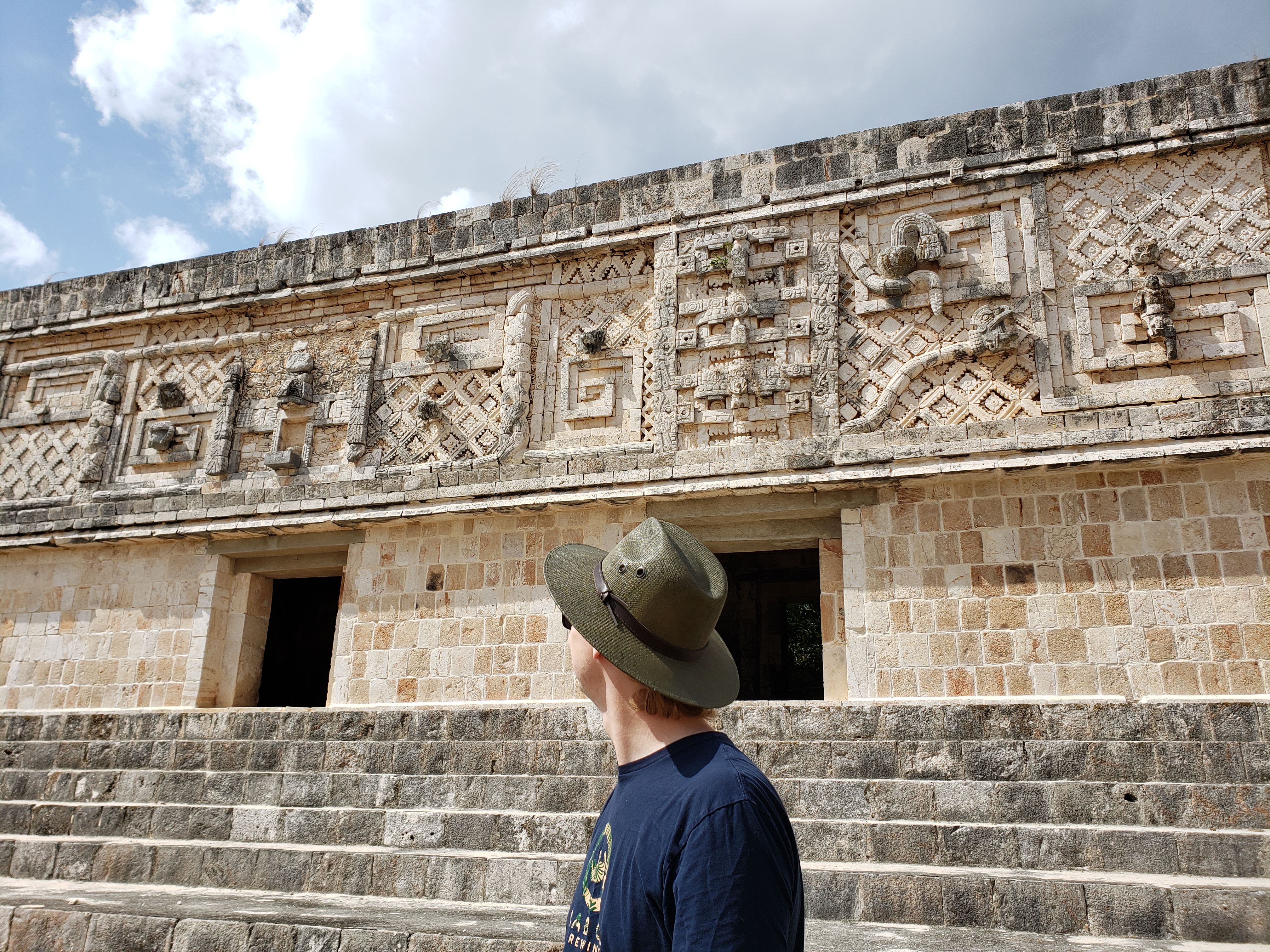 touring Uxmal, a Classical Mayan site in Mexico. Will is turning towards a set of stairs leading to a building with intricate stone carvings depicting the Mayan rain god, Chaac, and other figures from Mayan cosmology.